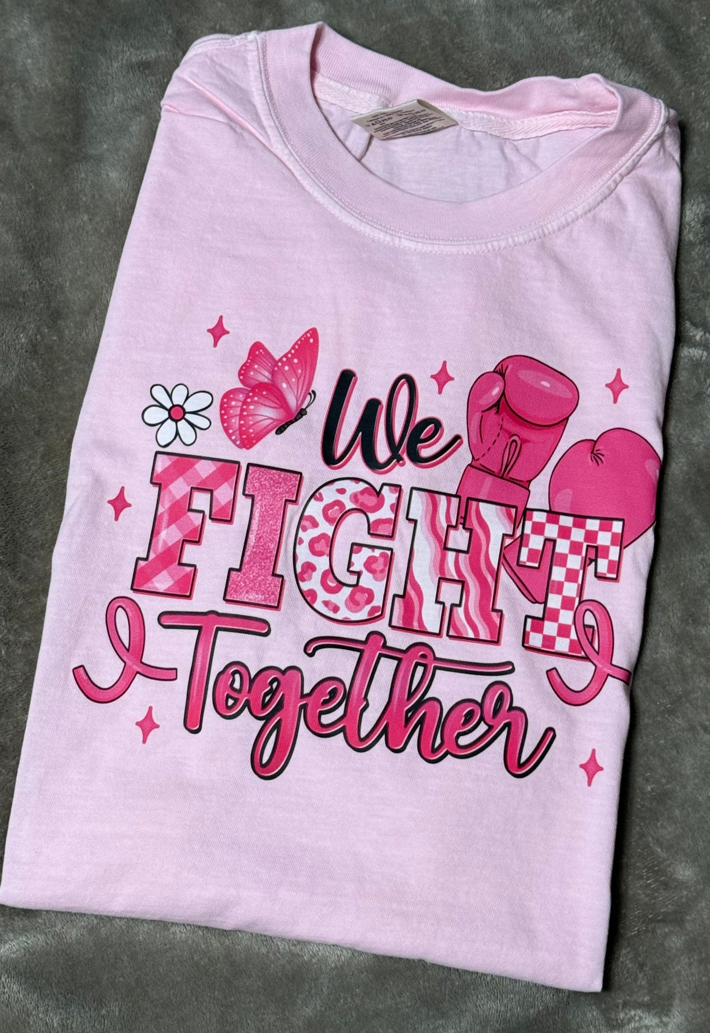 Pink t-shirt with 'We Fight Together' text and boxing glove graphics on a gray surface